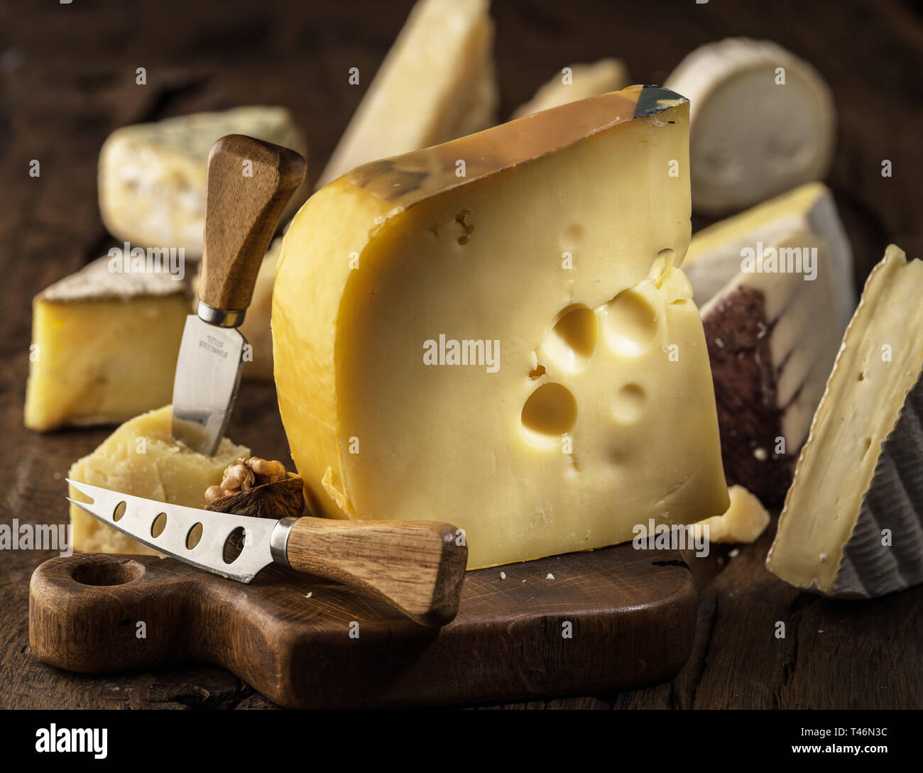 Parte de la leche de vaca Maasdam queso sobre la plancha de madera. Gama de quesos en el fondo. Foto de stock