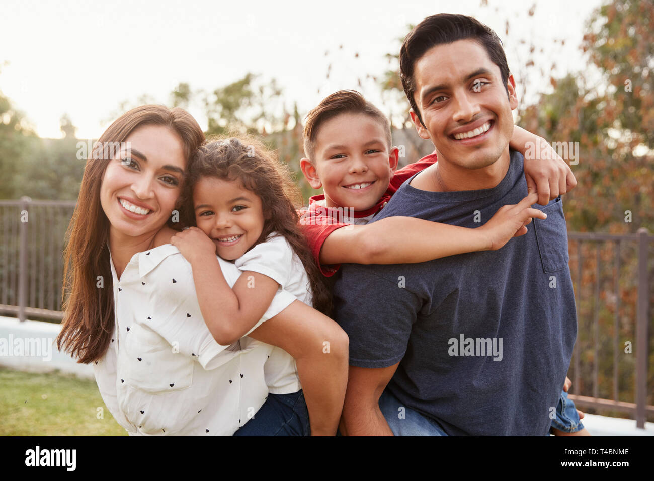 Los padres hispanos jóvenes piggyback a sus hijos en el parque, sonriendo a la cámara, se enfocan en primer plano Foto de stock