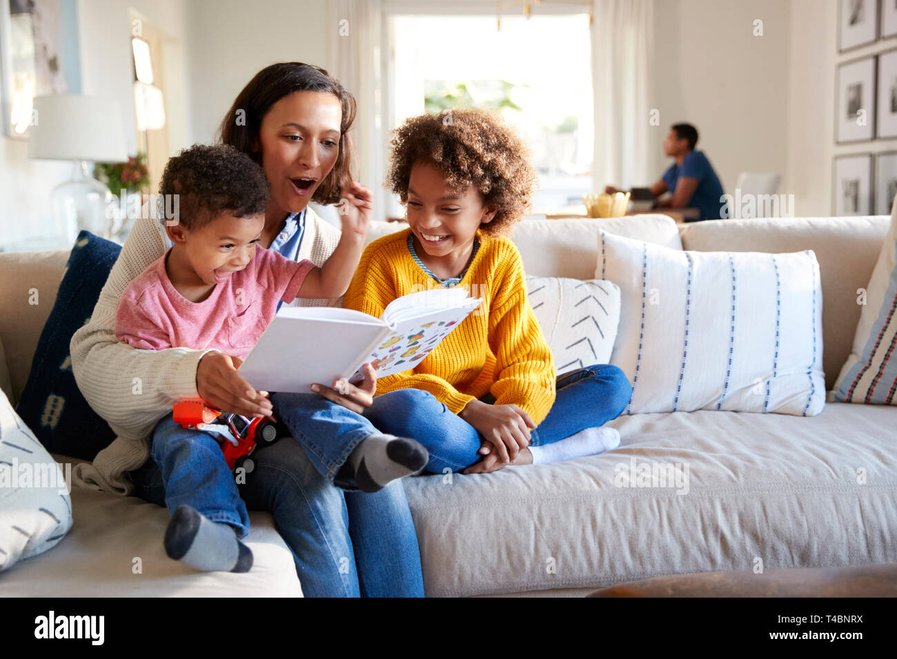 Cerca de la joven madre sentada en un sofá en el salón leyendo un libro a sus dos hijos, el padre estaba sentado en una mesa al fondo, se enfocan en primer plano Foto de stock