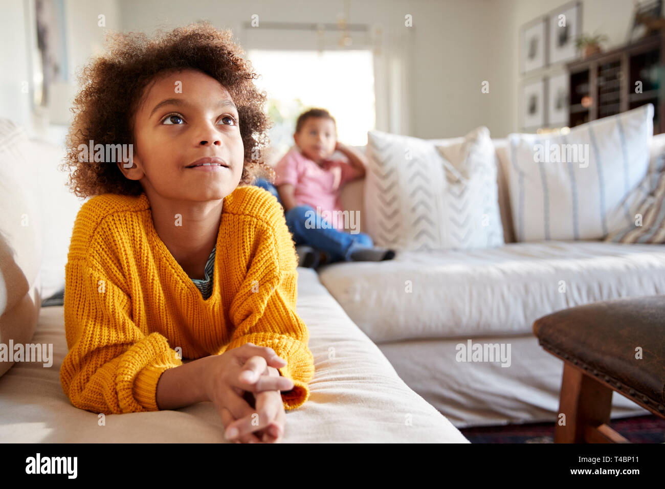Cierre de pre-teen girl tumbado en el sofá viendo la TV en el salón, su hermano sentado en el fondo, se enfocan en primer plano Foto de stock