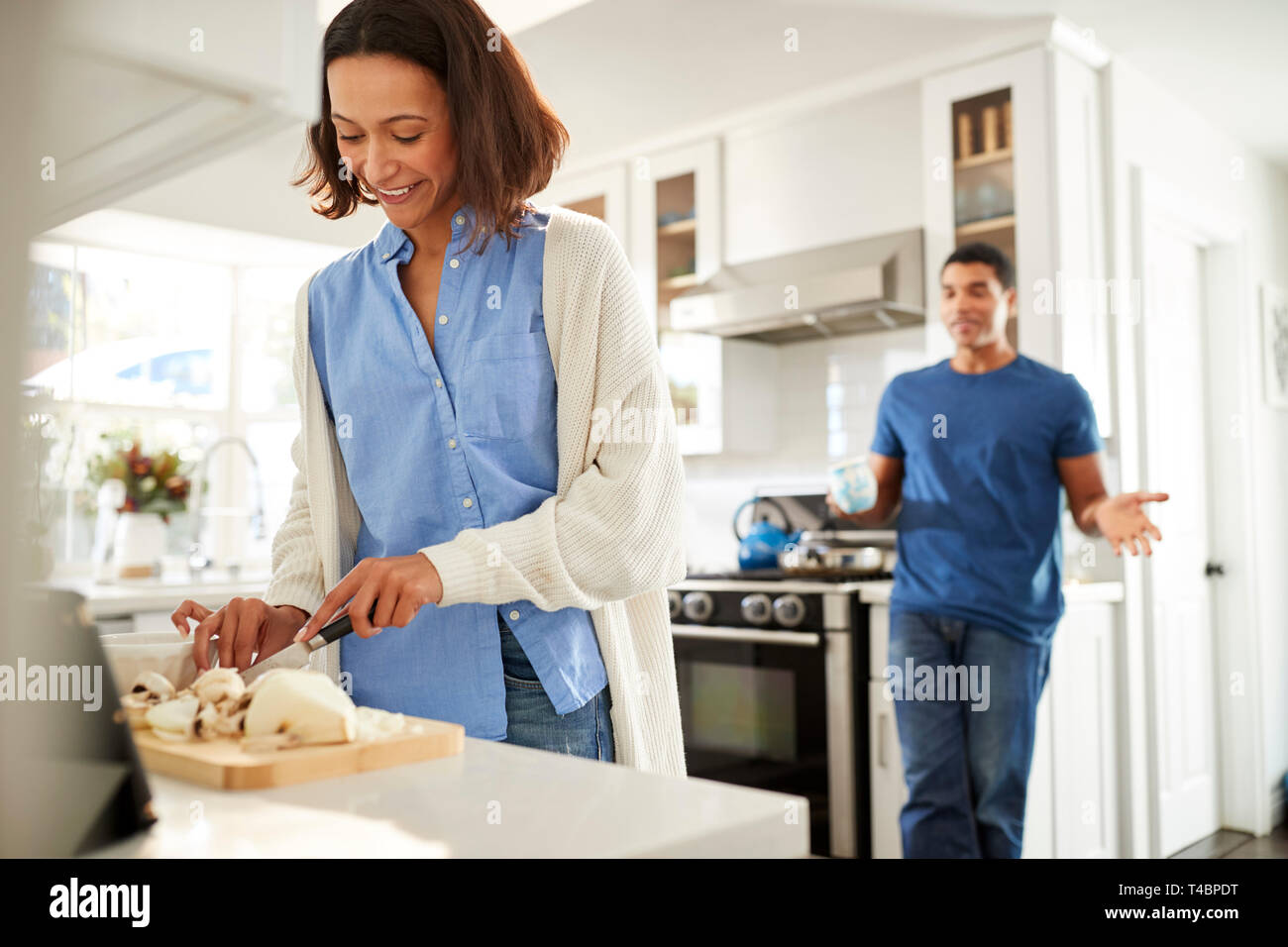 Joven mujer de raza mixta de pie en la cocina preparando la comida, su compañera de pie detrás de hablar, centrarse en primer plano Foto de stock