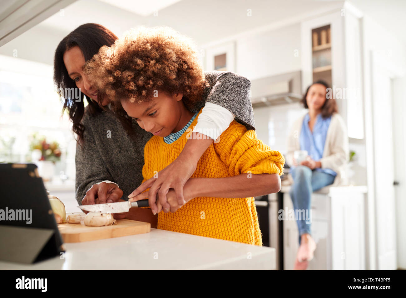 Abuela ayudando a su nieta a preparar la comida en la cocina, la Madre sentada en el fondo, se enfocan en primer plano Foto de stock