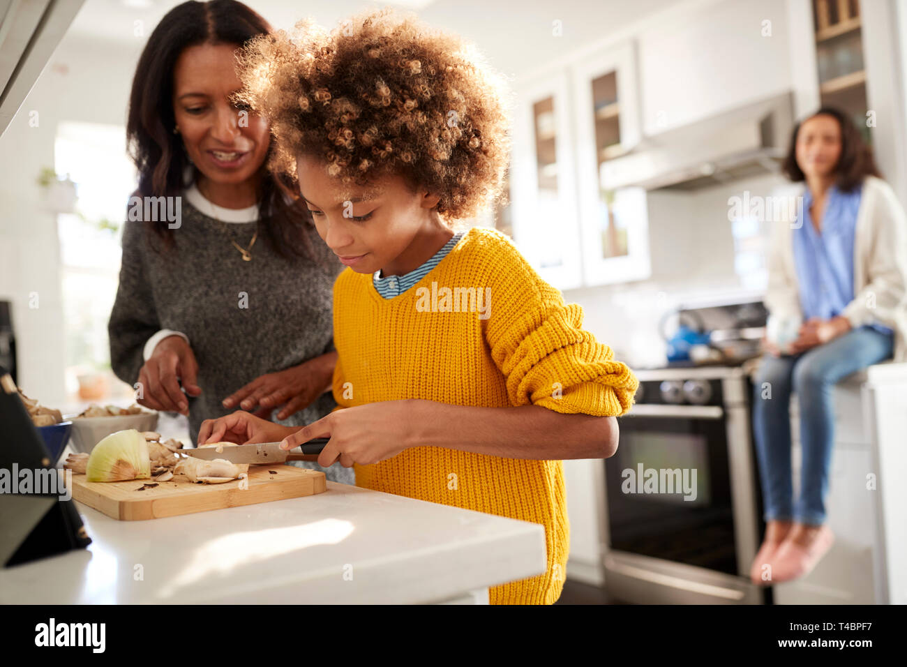 Abuela ayudando a su nieta a preparar la comida en la cocina, la Madre sentada en el fondo, se enfocan en primer plano Foto de stock