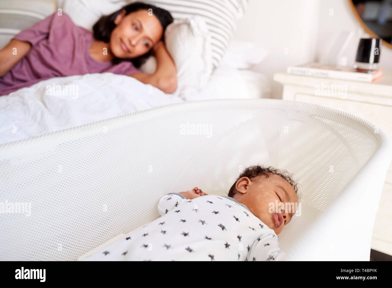 Cerca del adulto joven madre acostada en la cama mirando a su bebé de tres meses durmiendo en su cuna Foto de stock