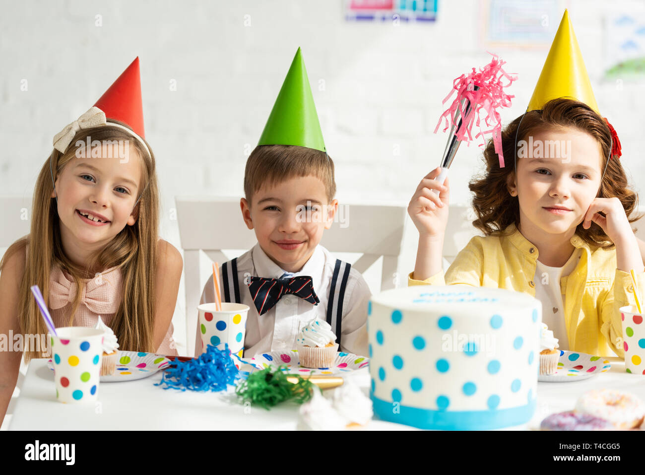 Los niños con gorros de fiesta sentado en la mesa y mirando a la cámara  durante la fiesta de cumpleaños Fotografía de stock - Alamy