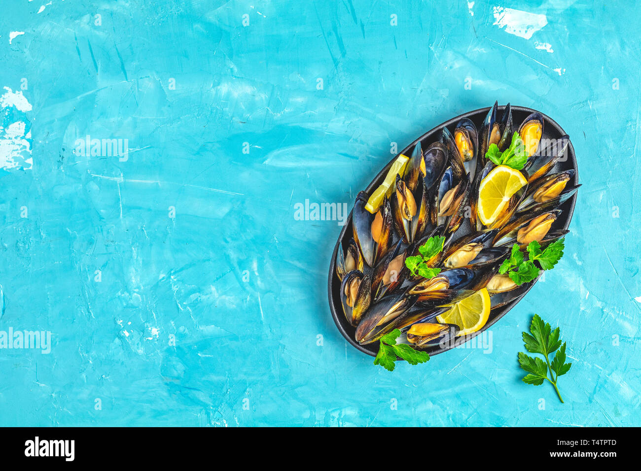 Mariscos mejillones con limón y perejil en placa metálica negra sobre la superficie de la mesa de hormigón de color azul, vista superior, copia espacio para que el texto Foto de stock