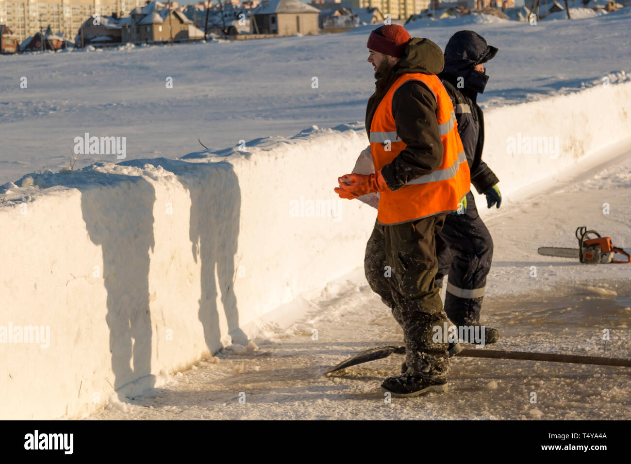 Un trabajador en un chaleco reflectante naranja lleva un bloque de hielo desde el orificio en su mano. Foto de stock