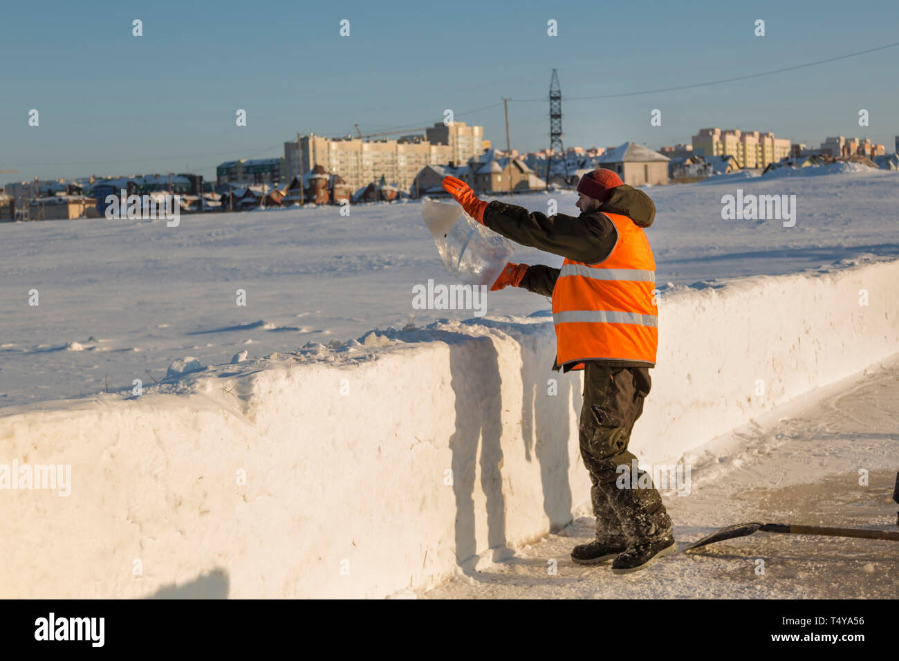 Un trabajador en un chaleco reflectante naranja lanza un bloque de hielo sobre un montículo de nieve. Foto de stock