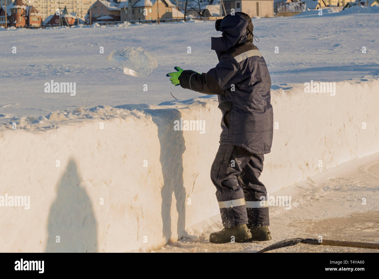 Trabajador en una chaqueta azul con un capó lanza un bloque de hielo de un montículo de nieve Foto de stock
