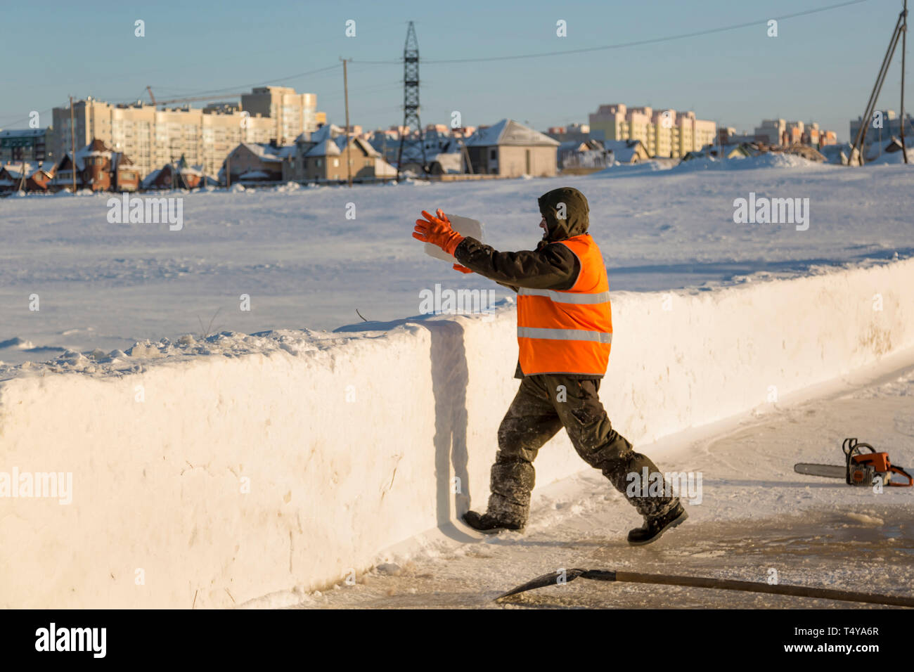 Un trabajador en un chaleco reflectante naranja lanza un bloque de hielo sobre un montículo de nieve. Foto de stock