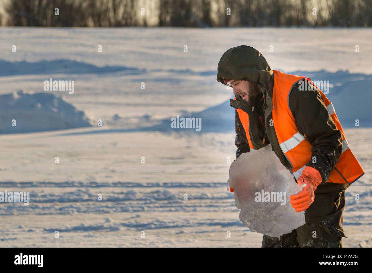Un trabajador en un chaleco reflectante naranja lleva un bloque de hielo desde el orificio en su mano. Foto de stock