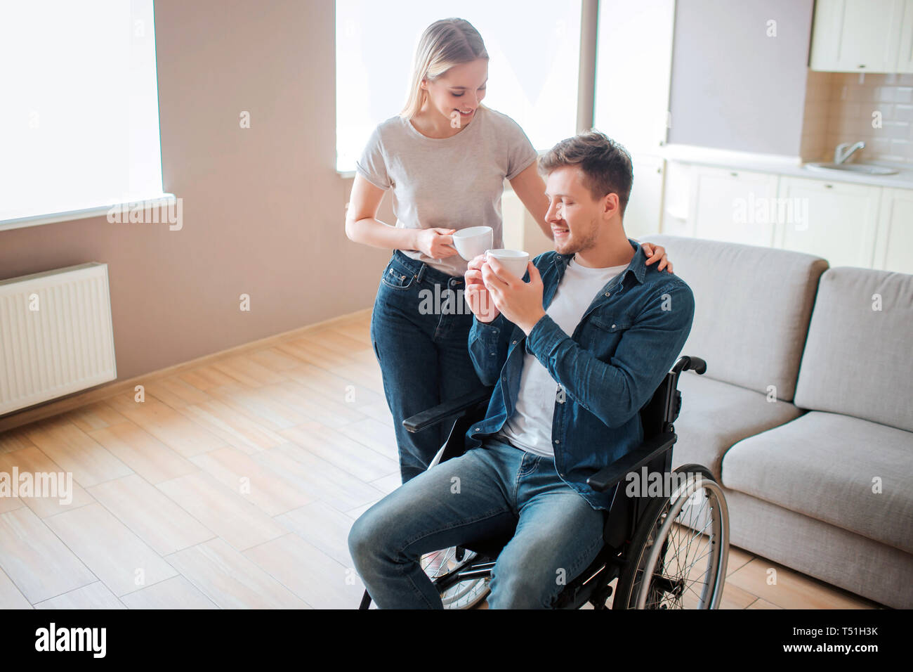 Joven con discapacidad sentarse en una silla de ruedas. Las personas con necesidades especiales. Celebración de una taza de café junto con mi novia. Ella stand aparte y mantenga la nad Foto de stock