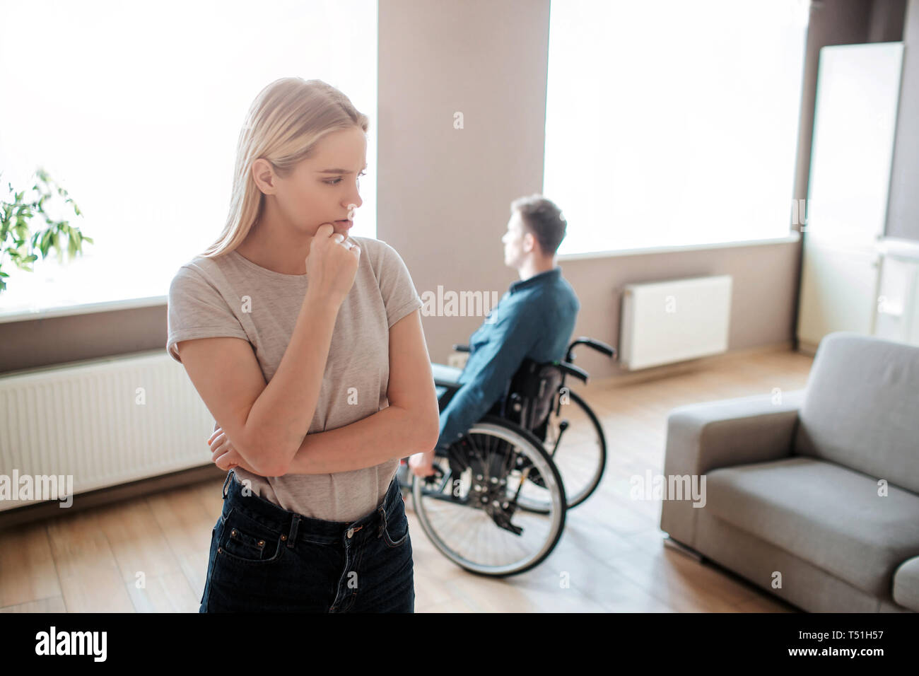 Joven sentarse con necesidades especiales sentarse en silla de ruedas y mirar en la ventana. Mujer de pie delante y pensar. Discutir y quirrel. Par malestar e infeliz Foto de stock