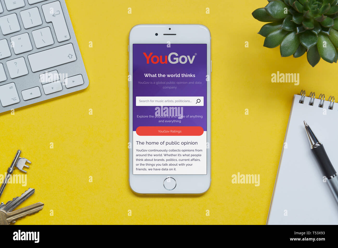Un iPhone que muestra el sitio web de YouGov descansa sobre un fondo amarillo tabla con un teclado, teclas, bloc de notas y Planta (uso Editorial solamente). Foto de stock