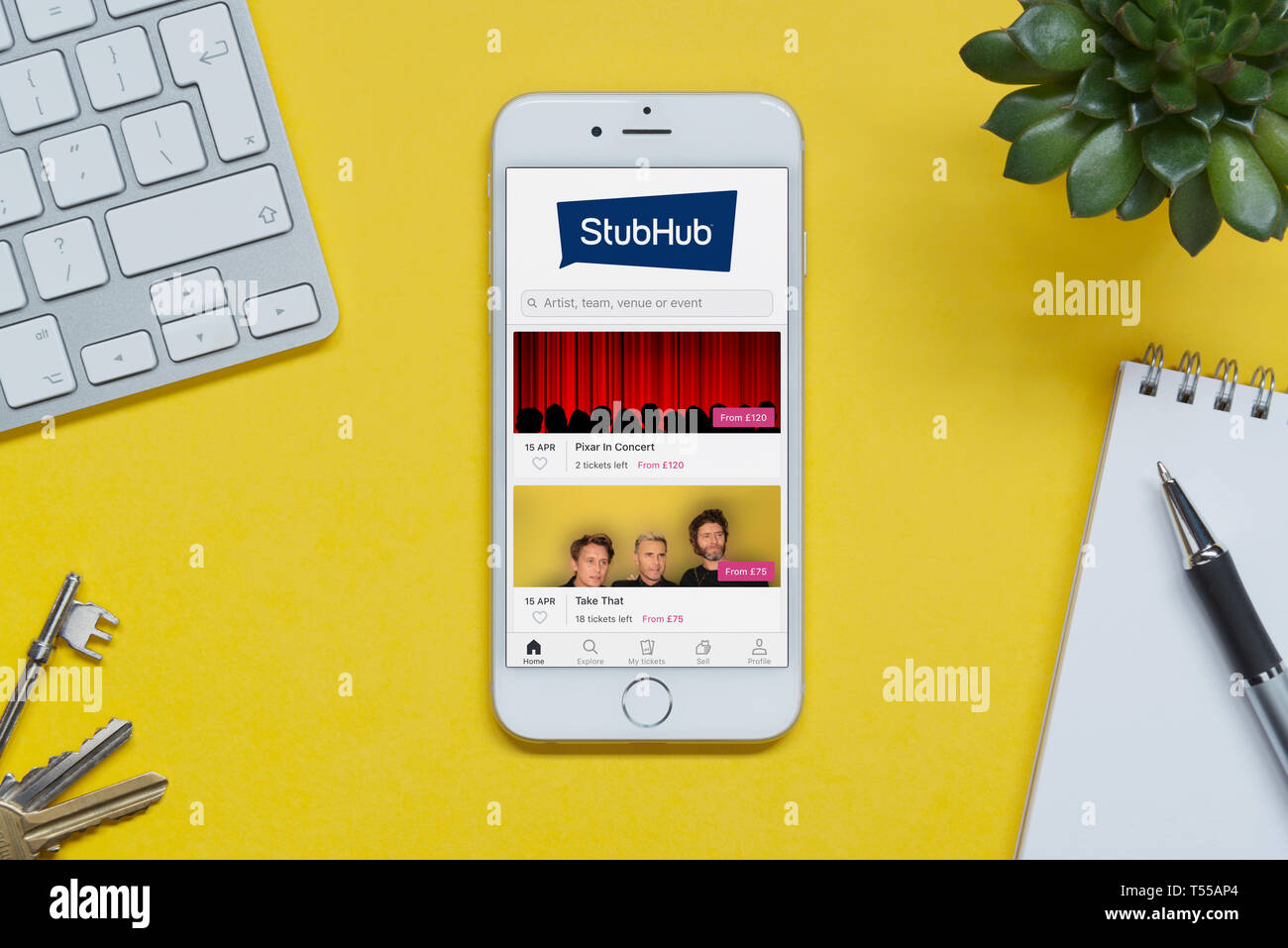 Un iPhone que muestra el sitio StubHub descansa sobre un fondo amarillo tabla con un teclado, teclas, bloc de notas y Planta (uso Editorial solamente). Foto de stock