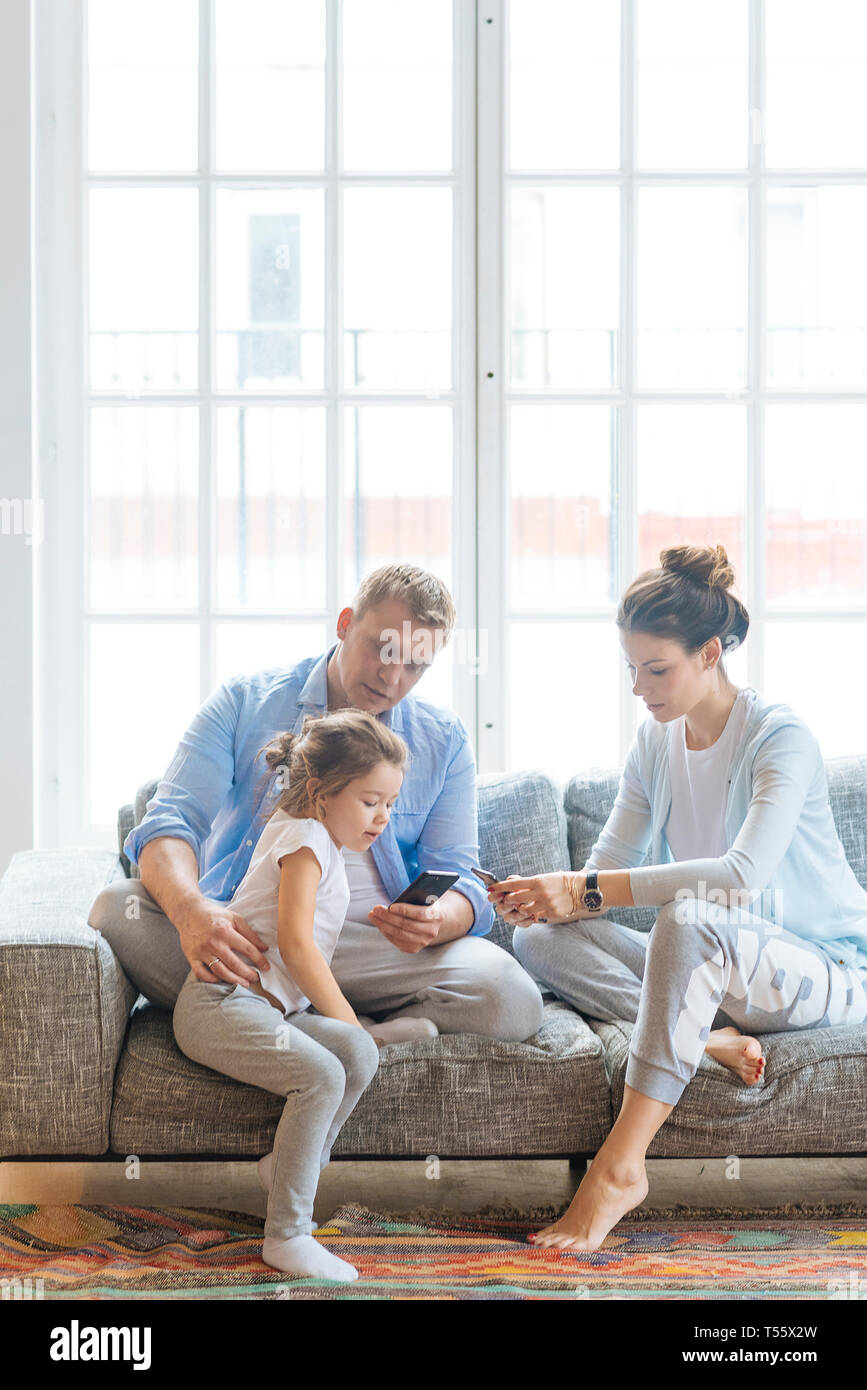 Padres que tengan teléfonos inteligentes en el sofá con su hija Foto de stock