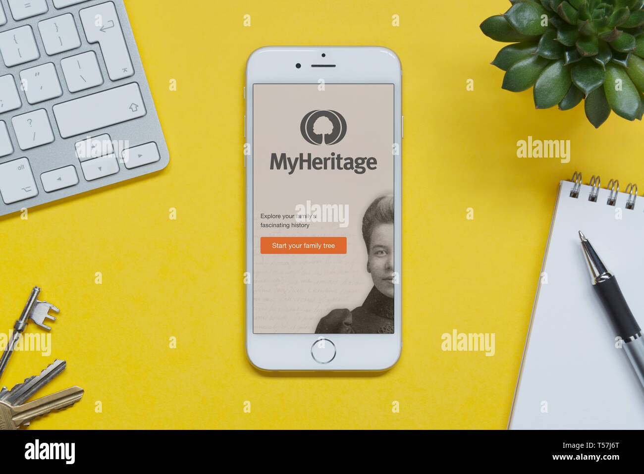 Un iPhone que muestra el sitio web de mi herencia descansa sobre un fondo amarillo tabla con un teclado, teclas, bloc de notas y Planta (uso Editorial solamente). Foto de stock