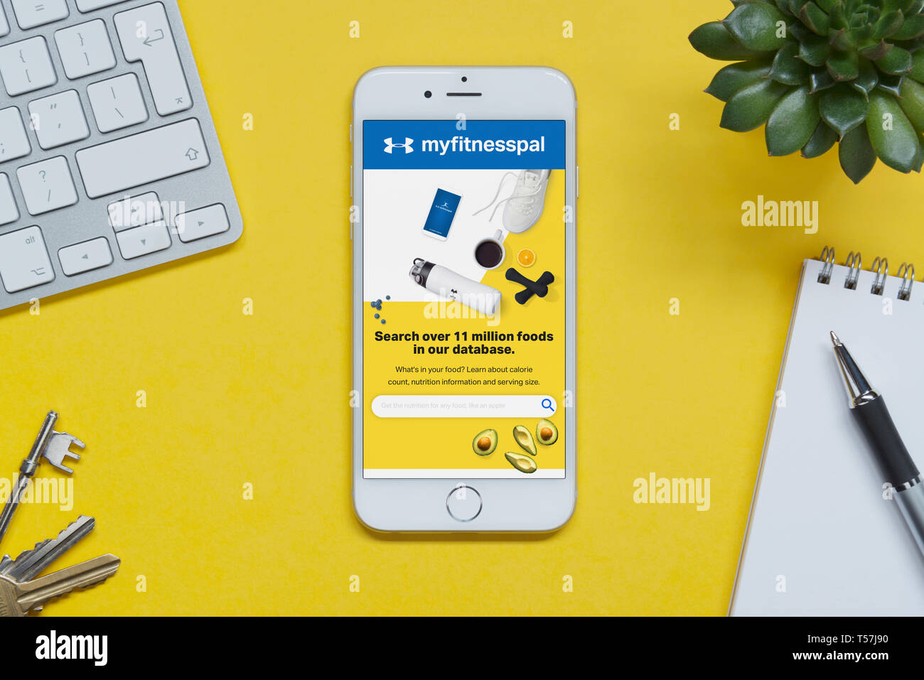 Un iPhone que muestra el sitio web myfitnesspal descansa sobre un fondo amarillo tabla con un teclado, teclas, bloc de notas y Planta (uso Editorial solamente). Foto de stock