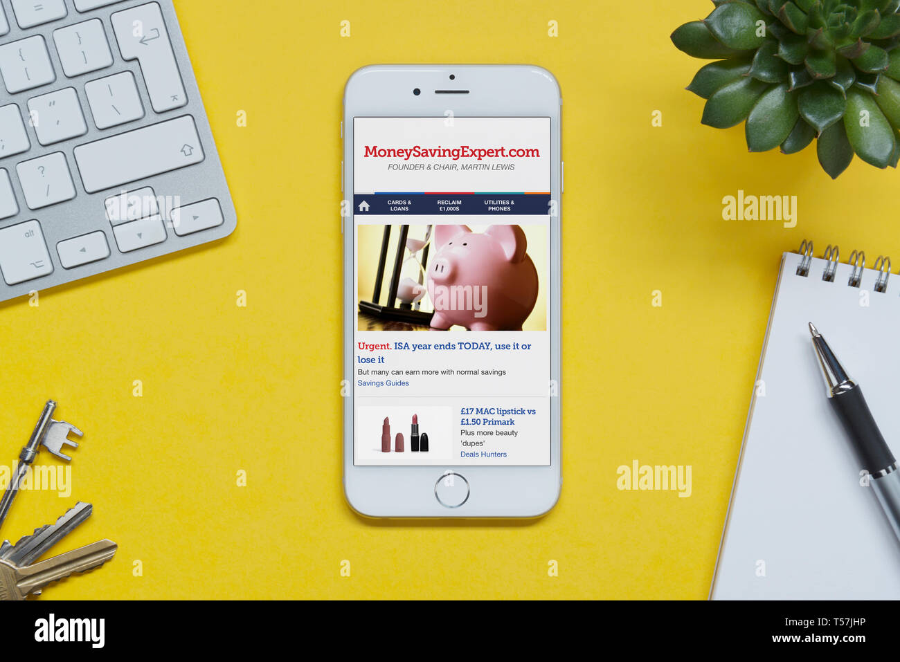 Un iPhone que muestra el sitio web de expertos ahorrar dinero descansa sobre un fondo amarillo tabla con un teclado, teclas, bloc de notas y Planta (uso Editorial solamente). Foto de stock
