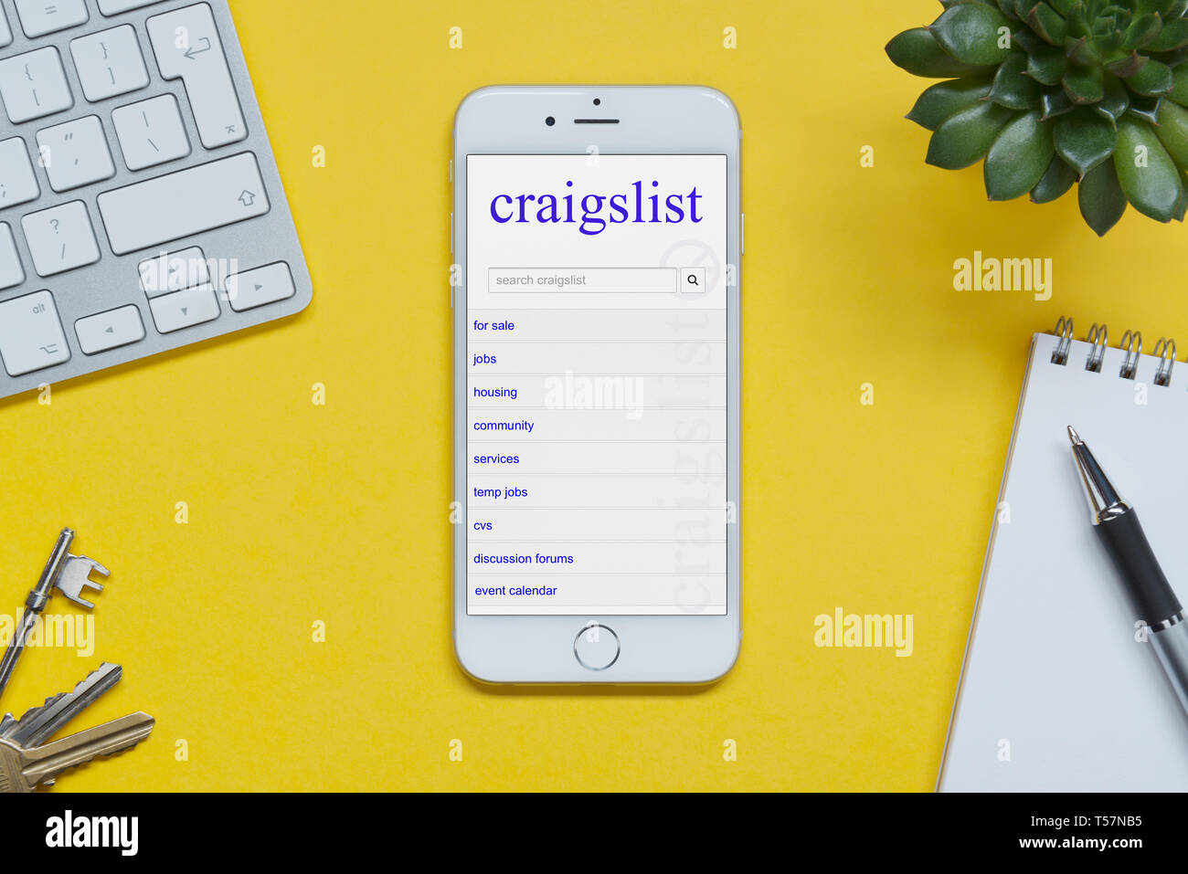 Un iPhone que muestra el sitio web Craigslist descansa sobre un fondo amarillo tabla con un teclado, teclas, bloc de notas y Planta (uso Editorial solamente). Foto de stock