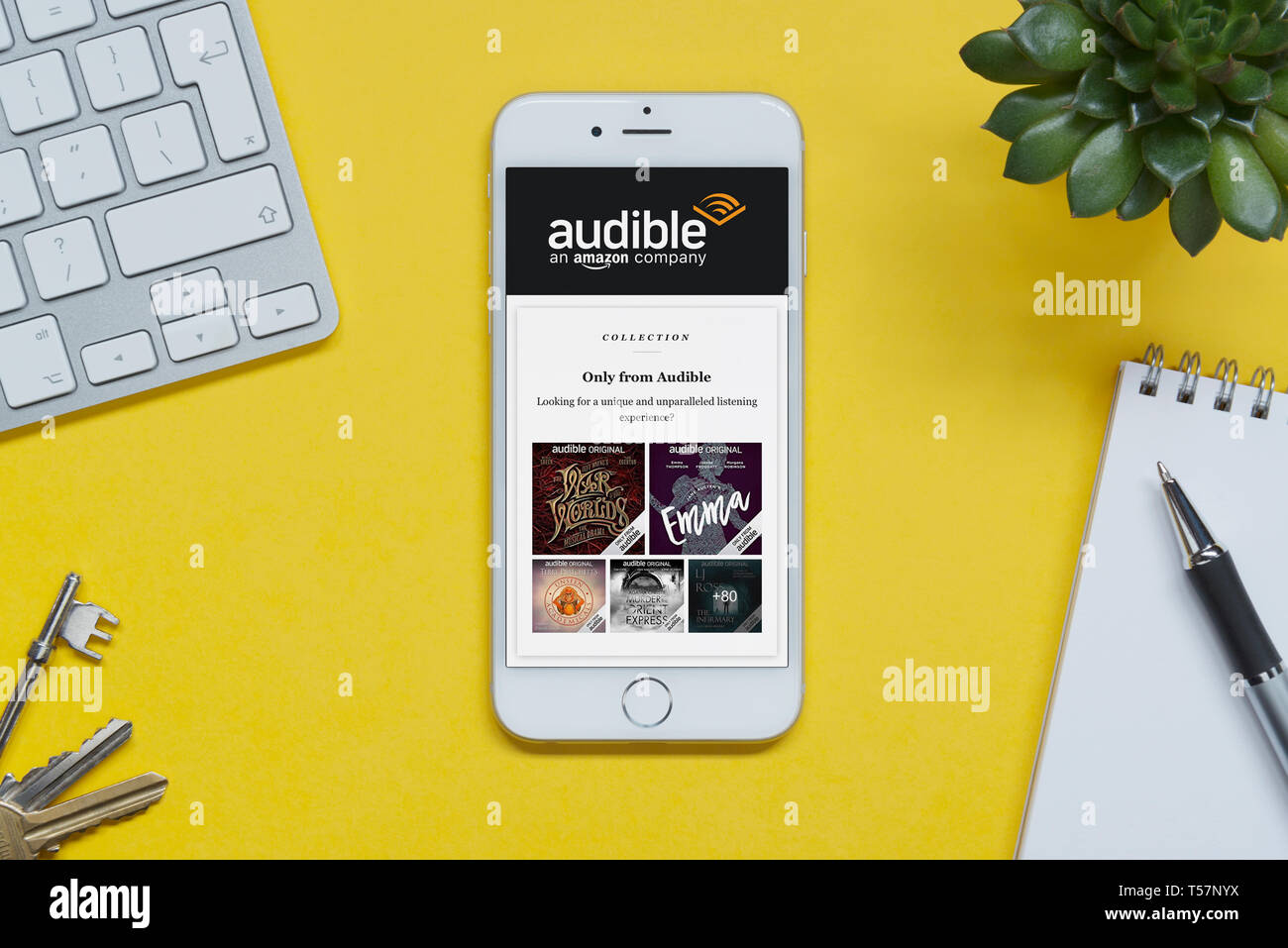 Un iPhone que muestra la página web Audible descansa sobre un fondo amarillo tabla con un teclado, teclas, bloc de notas y Planta (uso Editorial solamente). Foto de stock