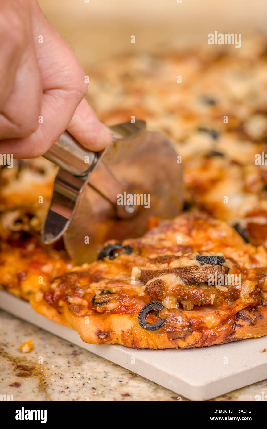 El hombre cortando un trozo de carne y veggie pizza casera con ingredientes de salchichas, salchichones, tocino canadiense, aceitunas negras, champiñones, queso mozzarella, Foto de stock