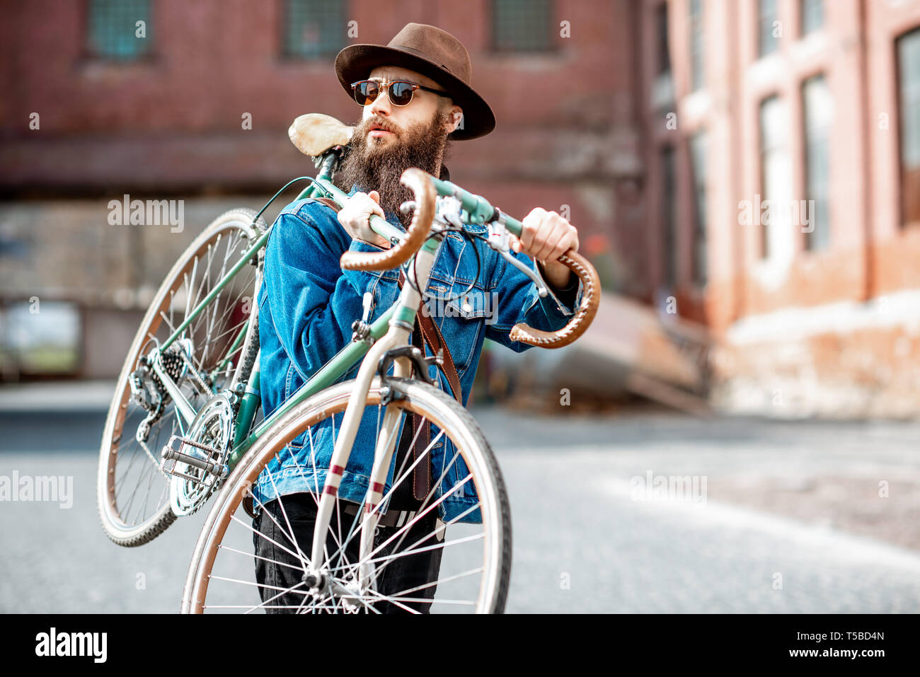Retrato de un estilo HIPSTER barbudo vestida elegantemente con sombrero y chaqueta retro llevando su bicicleta en el contexto urbano Foto de stock