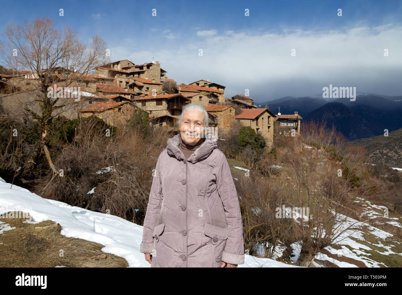 Una anciana mujer goza de un paisaje en un altiplano auténtico pueblo catalán con casas de piedra cuando caminaba por un sendero de montaña cubiertas de nieve que se derrite. Foto de stock