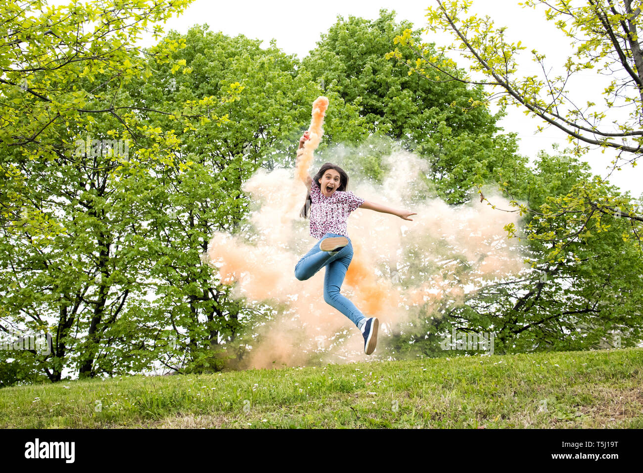 Ágil emocionado joven con un humo de color flare correr y saltar de alegría en un parque o jardín arbolado Foto de stock