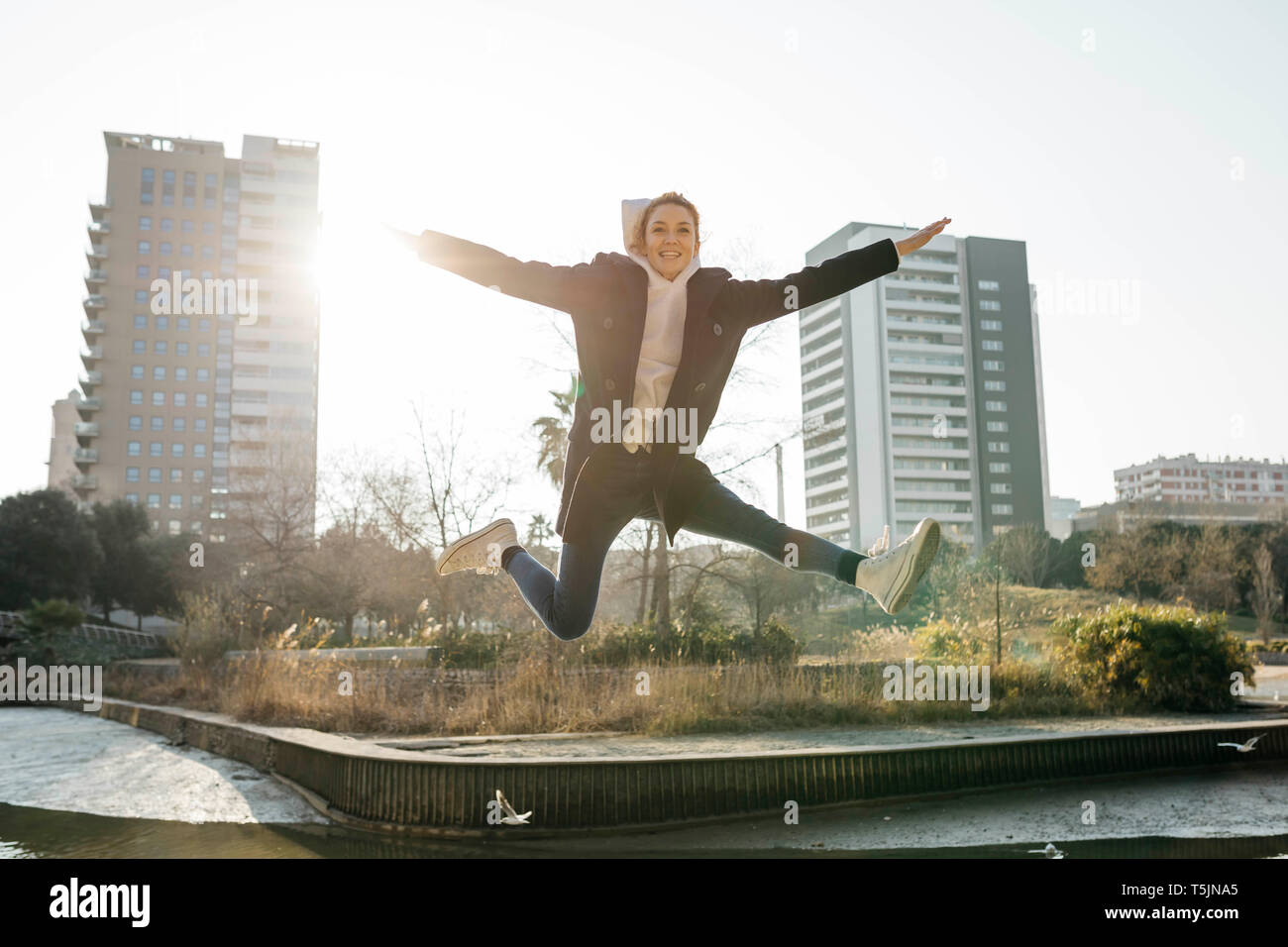 Mujer joven feliz saltando en un parque de la ciudad en segundo plano. Foto de stock