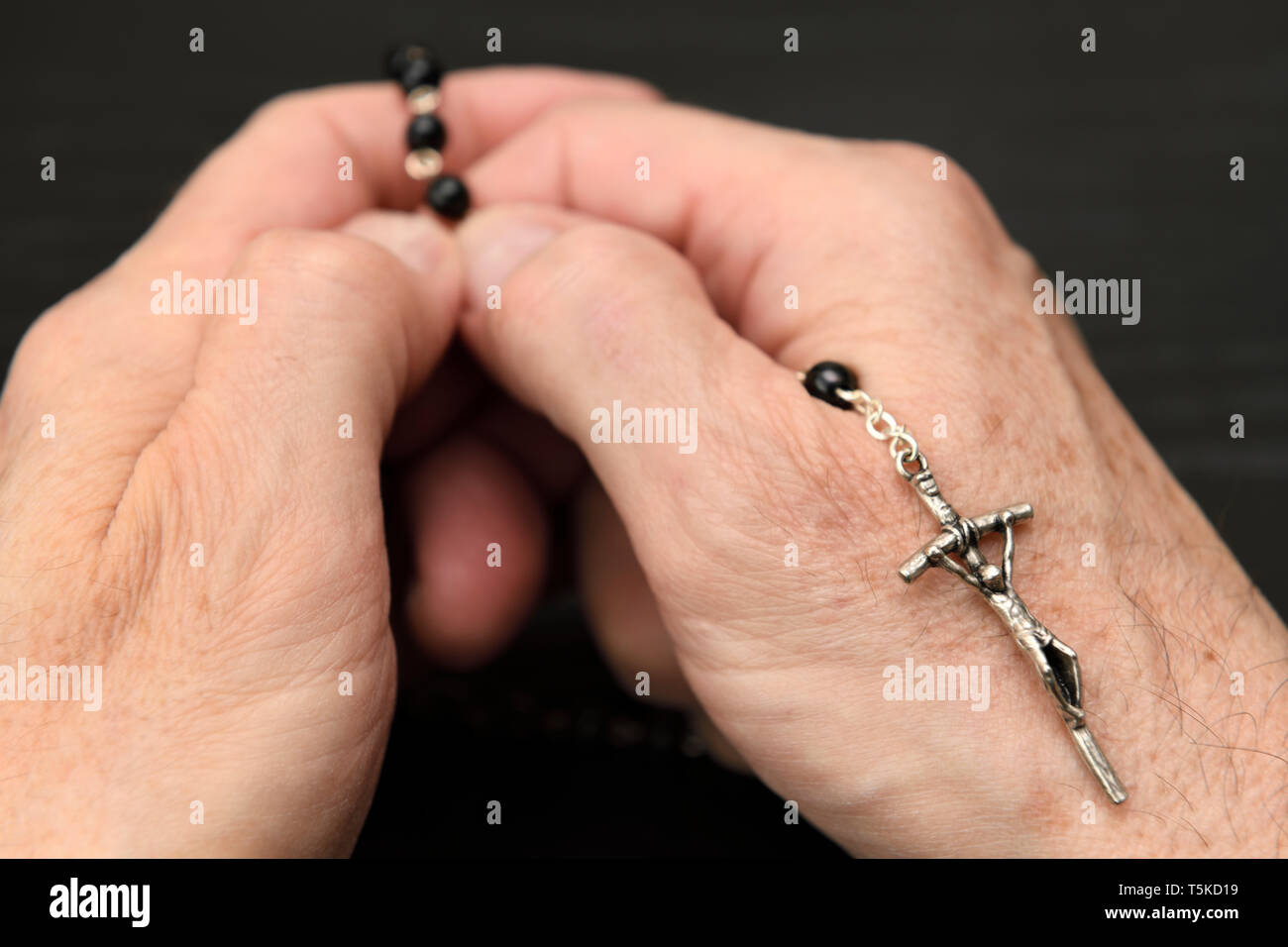 Manos de anciano meditando y orando en el negro de cordones de un santo rosario con crucifijo Foto de stock