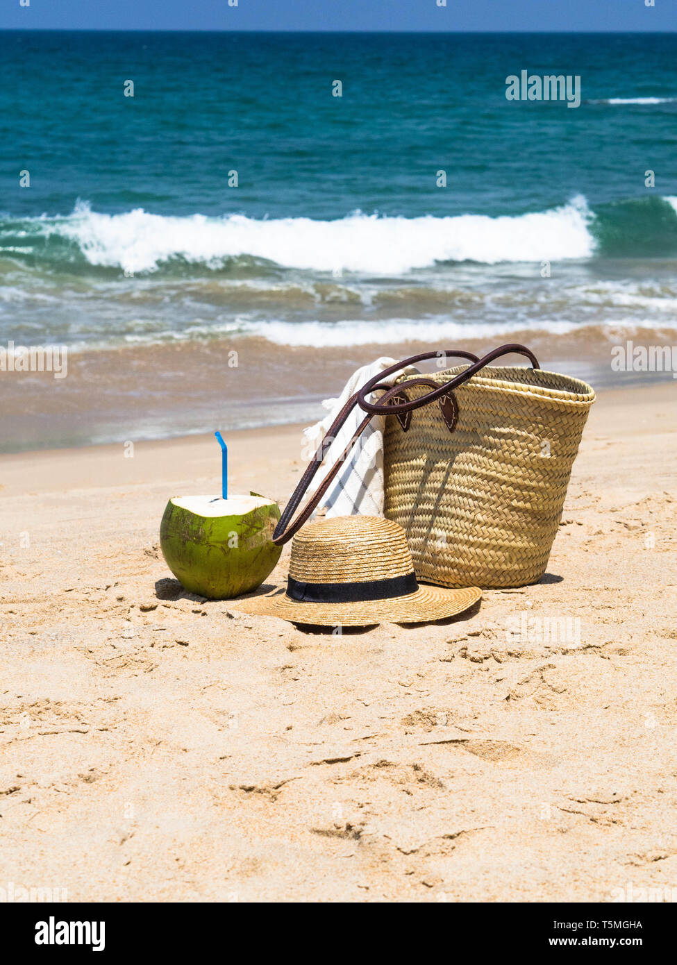 Un coco fresco y juvenil está listo para comer y una bolsa de paja y un  sombrero de paja de mujer en una playa de arena, frente a un mar azul.  Concepto