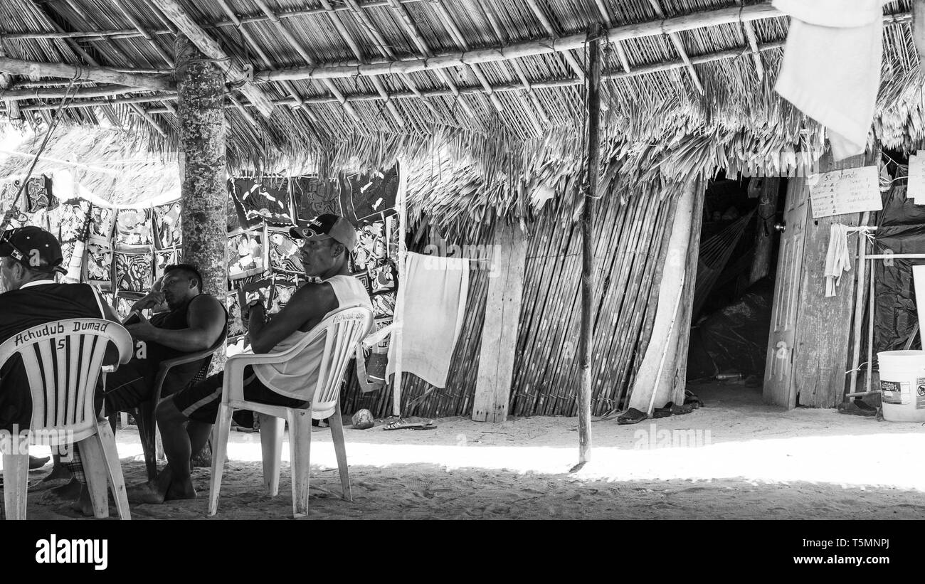 Guna (aka Kuna) miembros tribales comunal grande relajarse bajo una choza hecha con hojas de palmera, viendo como los turistas visitan su isla y comprar Molas Foto de stock