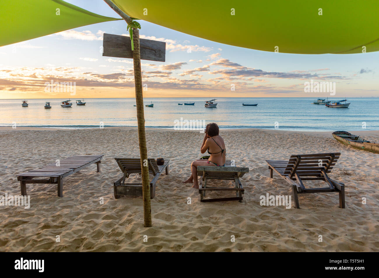 Bella mujer fotografiando atardecer en la playa idílica sentados con el mar y los barcos de fondo. La paz y la mente conceptual. Bahia, Brasil. Foto de stock