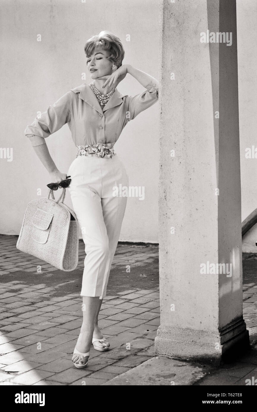1950 1960 Longitud total retrato Mujer vistiendo la camiseta ABAJO pantalones  capri sosteniendo una bolsa de paja gafas de sol posando POR COLUMNA -  f6552 DEB001 HARS CHIN B&W estilos sofisticados blusa