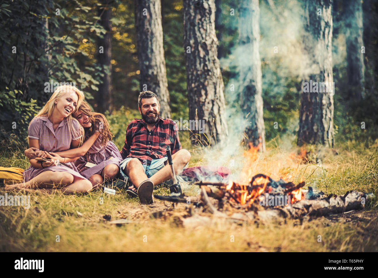 Hermosas chicas jóvenes vestidos retro en la lectura de libros en el campamento. Sonriente morena con cabello rizado rubio abrazando a su hermana mientras está sentado junto a la barba Foto de stock