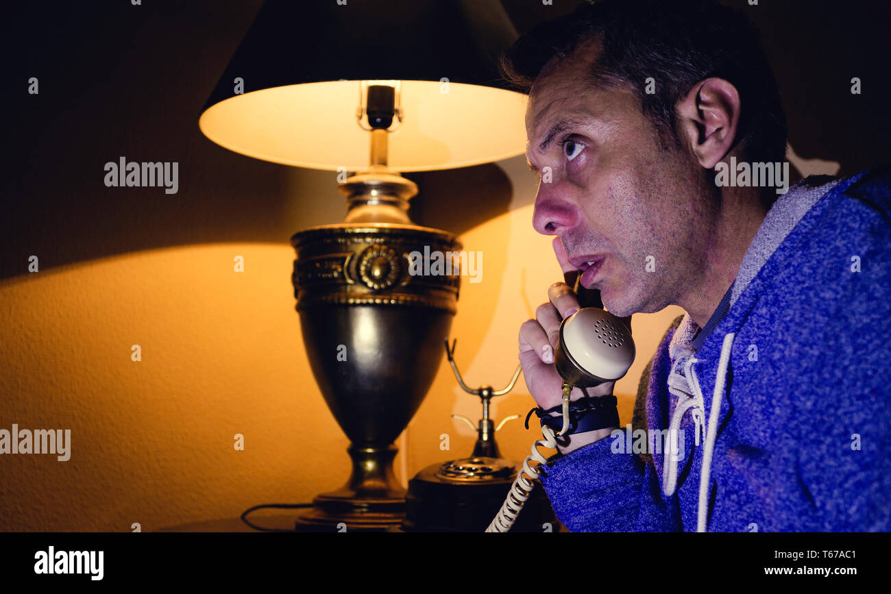 Retrato de un varón de mediana edad hablando con un teléfono vintage, con una lámpara de mesa encendida en el fondo Foto de stock