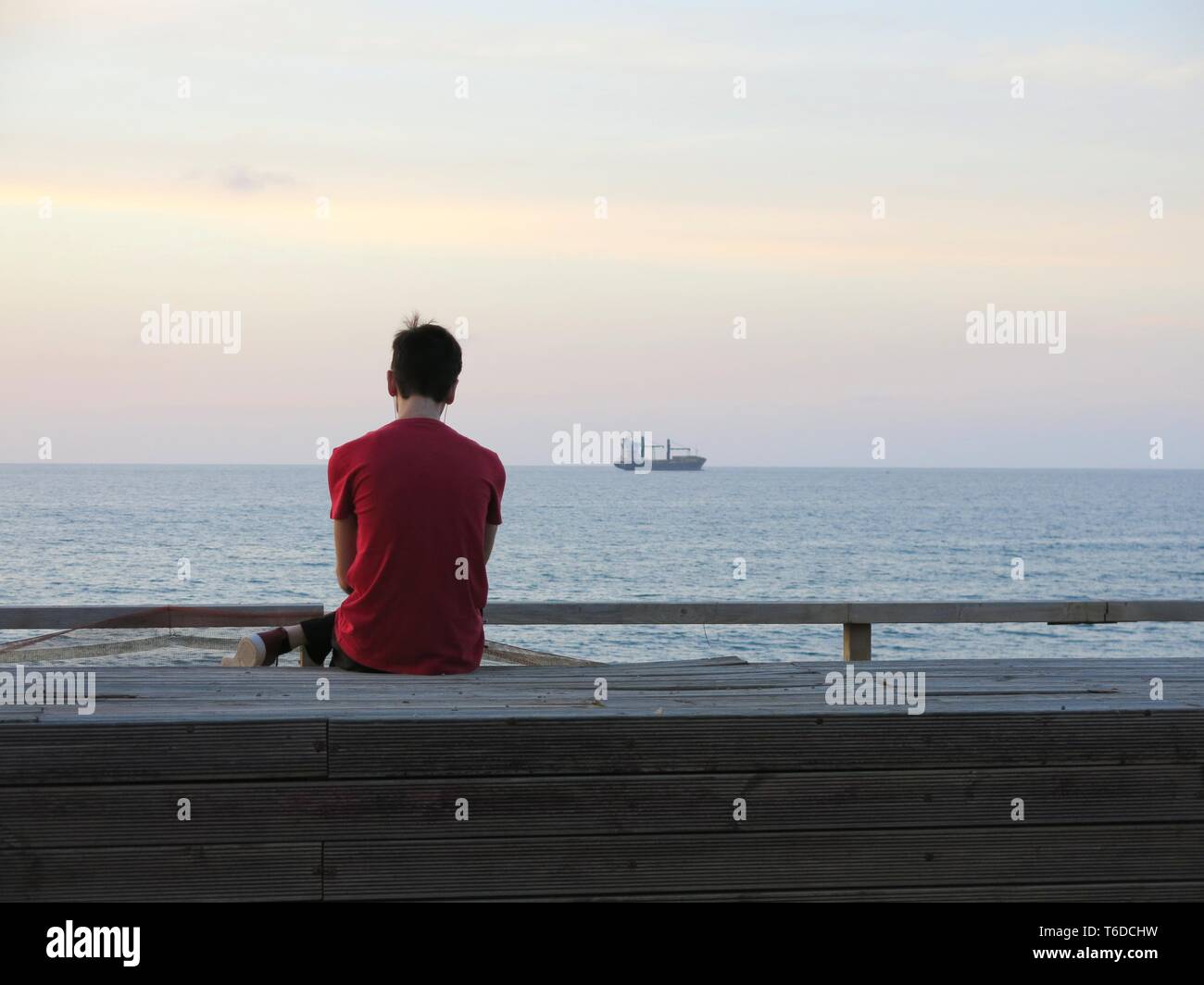 Joven sentada sola en el paseo mirando barco anclado frente a la costa, escuchando música ( concepto de soledad, relajación). Foto de stock