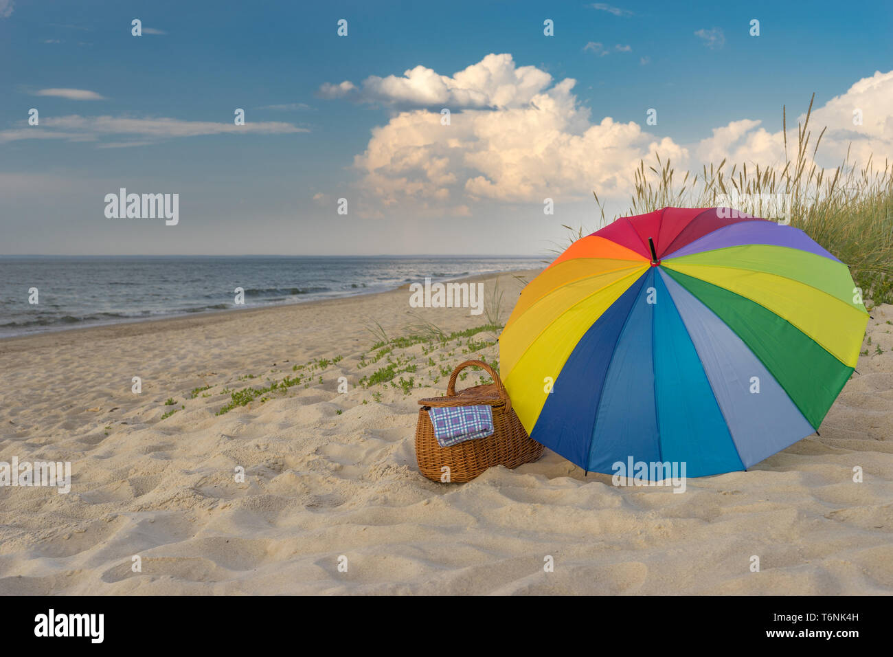 Paraguas multicolores y cesta de picnic frente a playa escénica y nubes, escapada de fin de semana, concepto Foto de stock