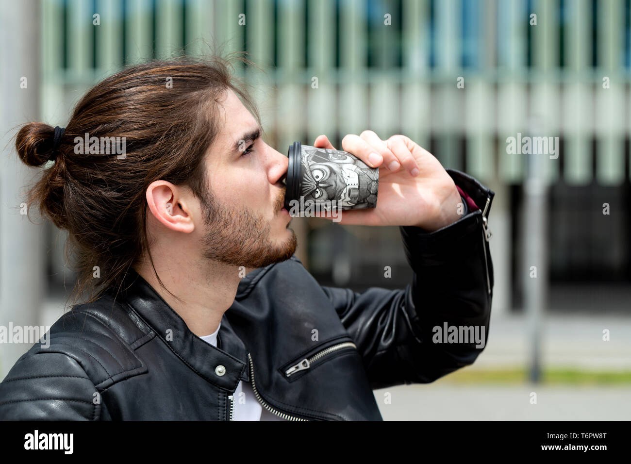 Retrato de un guapo de moda hipster hombre en una chaqueta de cuero negro bebiendo té o café de una taza de papel negro para llevar fuera. Foto de stock