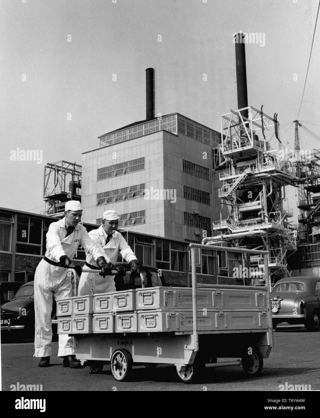 Ángulo de visión baja de dos trabajadores empujando cajas de elementos de combustible sobre una plataforma rodante, con el reactor número 2 en el fondo, Calder Hall, la central nuclear de Sellafield, Reino Unido, 1956. Imagen cortesía del Departamento de Energía de Estados Unidos. () Foto de stock