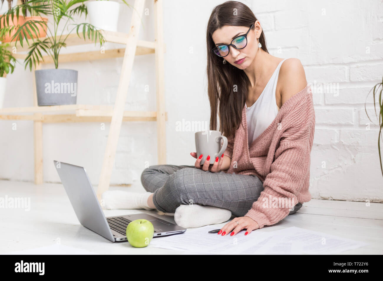 Mujer joven se sienta en el suelo, en un apartamento interior escandinavo con un portátil, estudiando derecho, una niña que trabaja en freelance, estudiante de aprendizaje a distancia Foto de stock