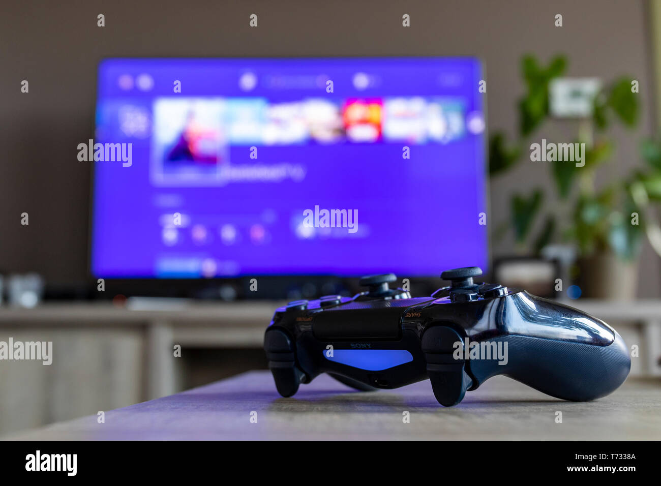 Un retrato de un controlador de PS4, que está encendida, delante de un televisor. La televisión es borrosa y muestra la pantalla de playstation home. Foto de stock