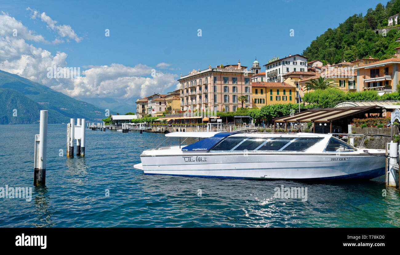 Un turista barco anclado en el puerto de Bellagio en el Lago de Como, con el Hotel Metropole y otros edificios coloridos más allá bajo un cielo azul brillante Foto de stock
