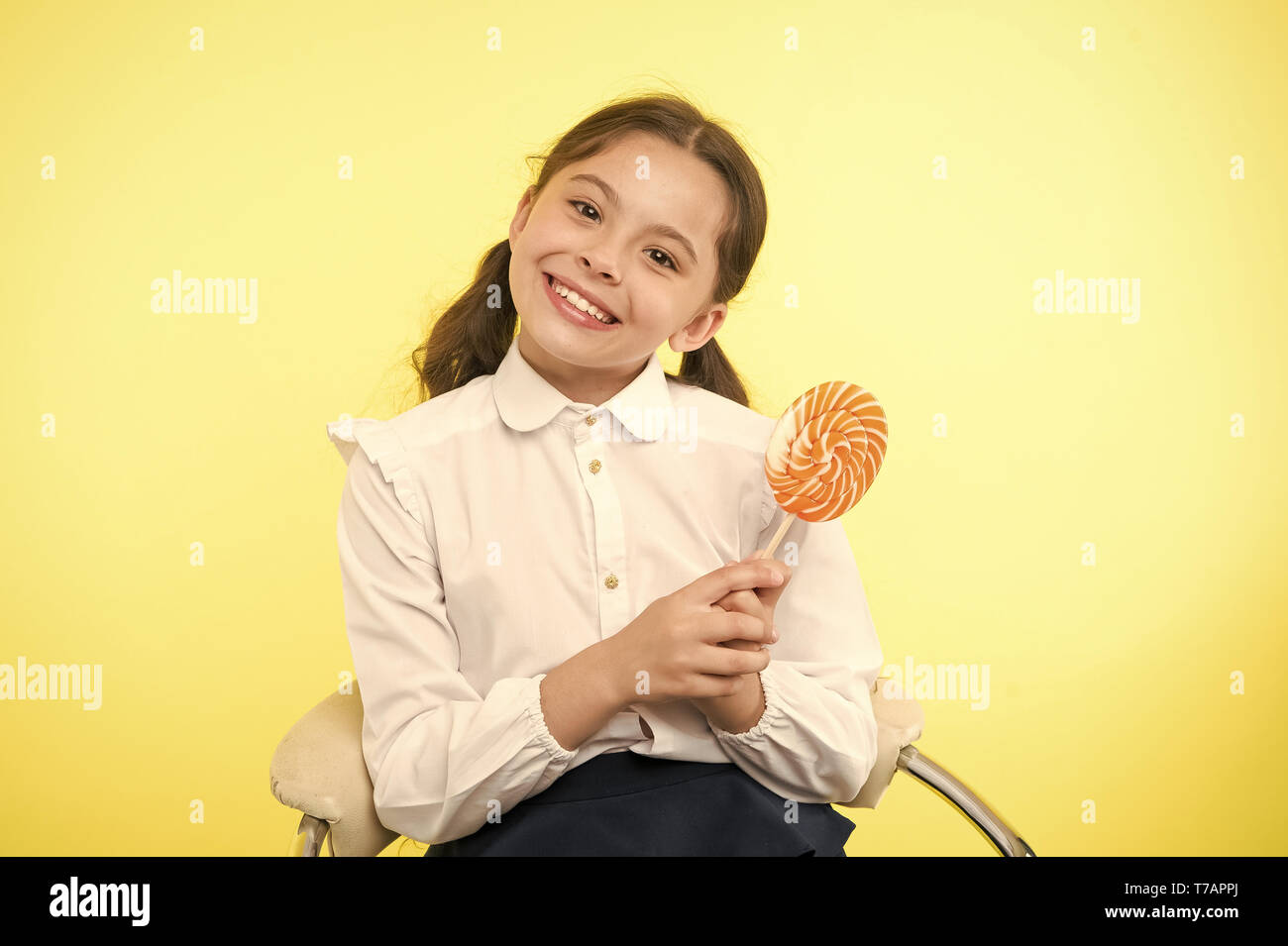 Divertirse con el caramelo. Chica chico lindo peinado ponytails comer dulces  lollipop. Dulces en porciones adecuadas ok. Alumna uniforme escolar le  gusta sweet lollipop fondo amarillo. Su sabor favorito Fotografía de stock -