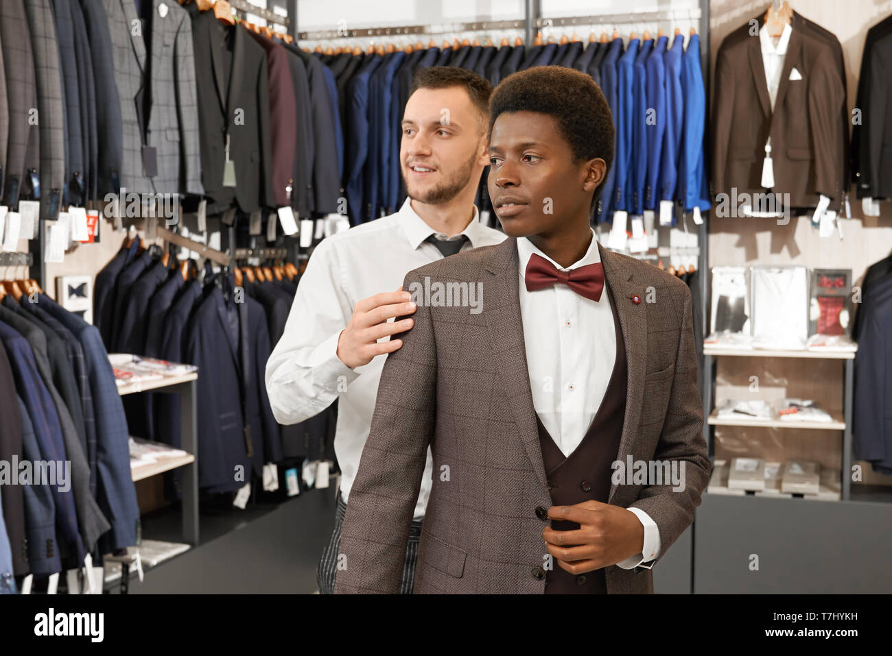 Assistant ayudando y eligiendo un traje para hombre africano en la elegante  tienda. Cliente en camisa blanca, chaleco, chaqueta y corbata roja y el  vendedor con barba de pie en el centro