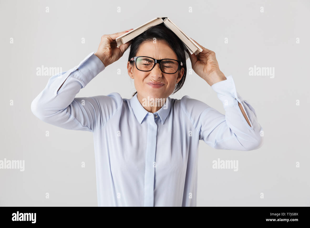 Foto de divertida trabajadora el uso de anteojos sosteniendo un libro sobre la cabeza en la oficina aislado sobre fondo blanco. Foto de stock