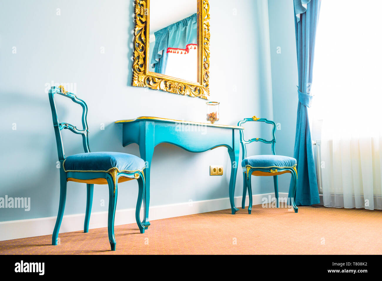 Vintage azul dos sillas y una mesa de madera junto a una pared con un  espejo, decorado con flores amarillas se arremolina en su marco. Cortinas  de color azul en el lado
