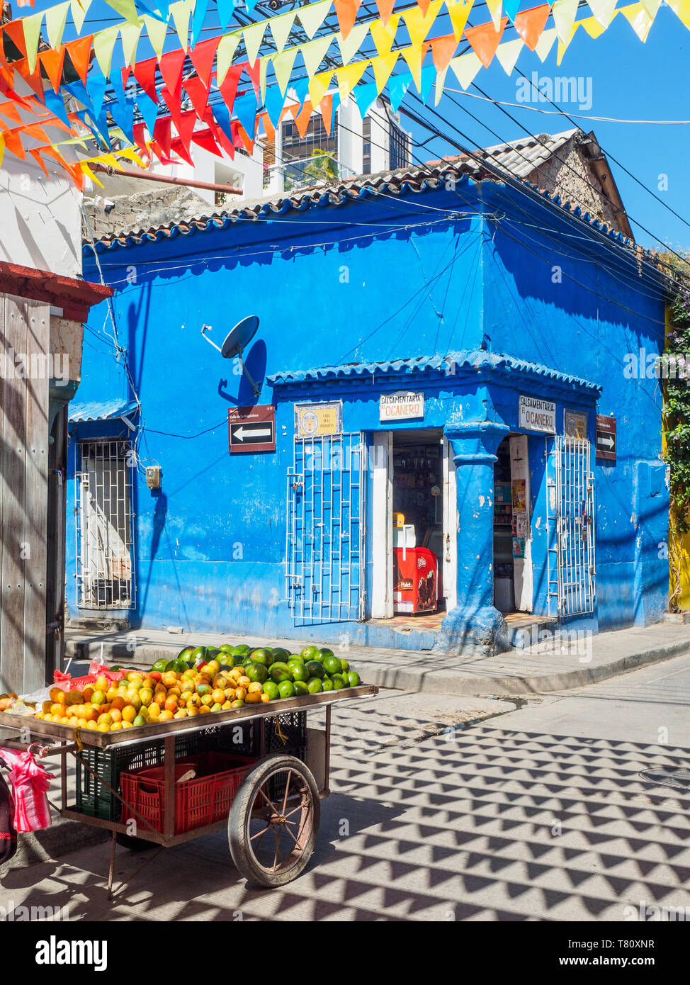 Coloridas banderas, edificio azul, y la cesta de frutas en la esquina de una calle en el barrio Getsemani, Cartagena, Colombia, Sur America Foto de stock