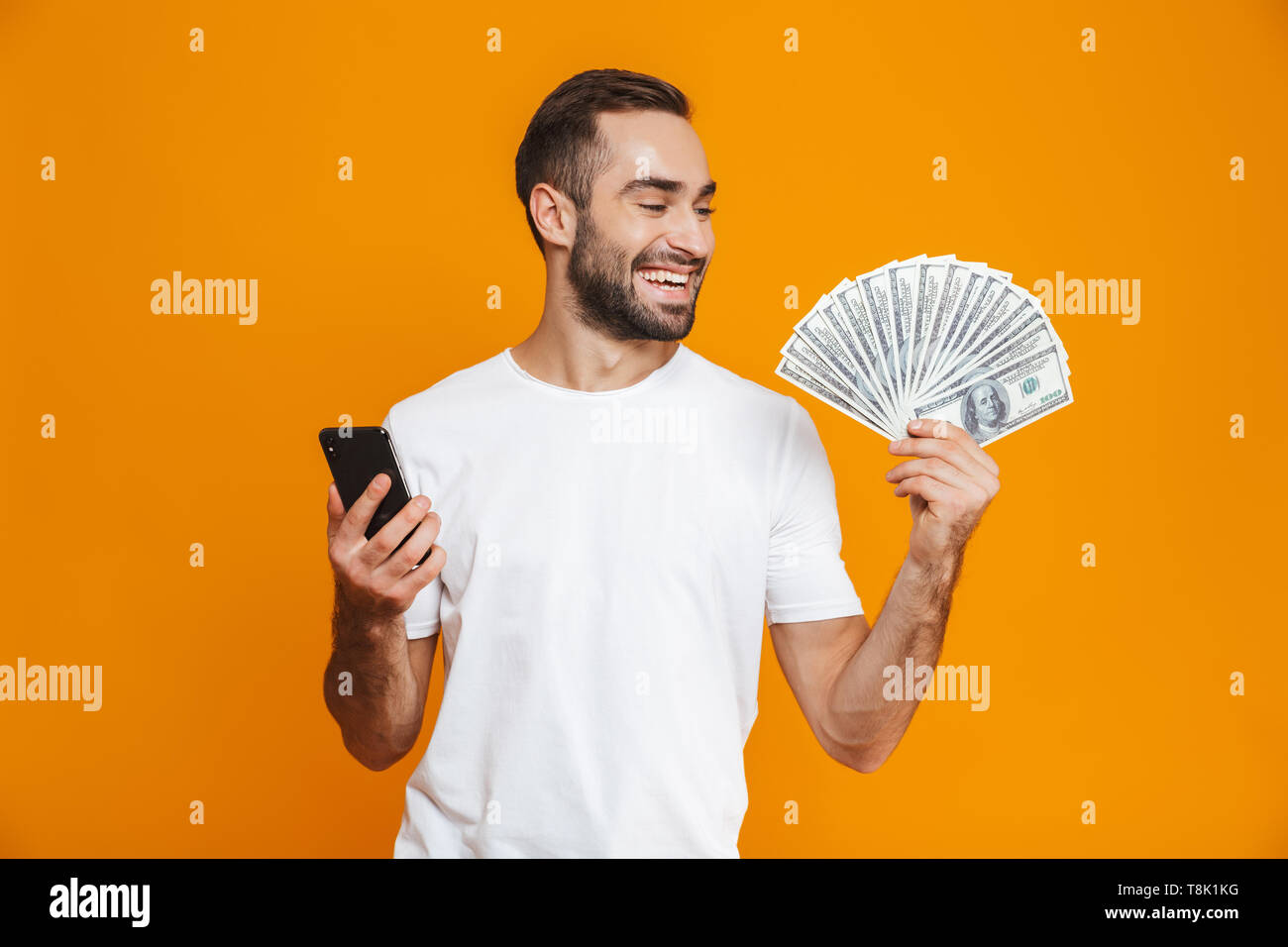 Foto del hombre sonriente 30s en ropa casual la celebración de teléfono celular y ventilador de dinero aislado sobre fondo amarillo Foto de stock